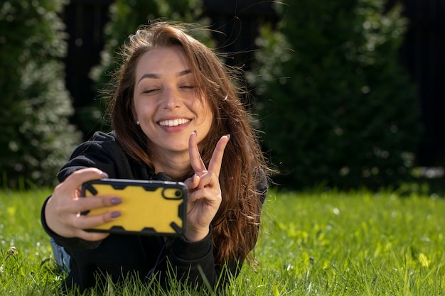 Фото Счастливая красивая женщина весело на открытом воздухе, делая селфи фото на мобильном телефоне со знаком мира, лежа на зеленой траве в парке