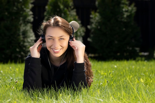 Happy beautiful woman in headphones lying on grass outdoors, listening to music