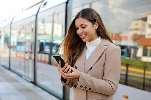 携帯電話の都市交通に関する更新情報をwebページに保持している幸せな美しい女。笑顔のビジネスウーマンがスマートフォンを介して電気交通機関に支払うオンラインチケットサービスに満足しています。