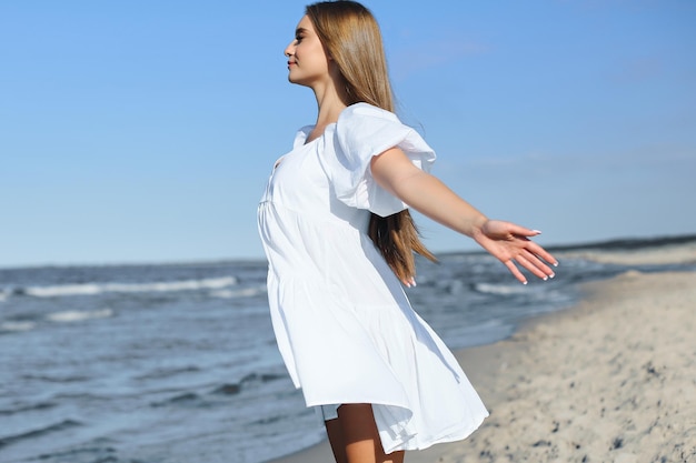 Happy, beautiful woman on the ocean beach standing in a white summer dress, open arms.