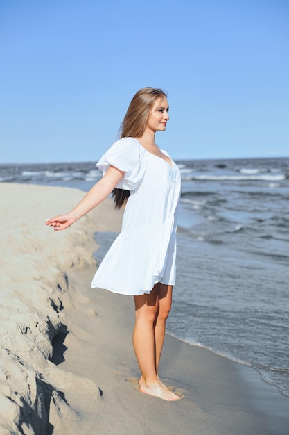 바다 해변에 있는 행복하고 아름다운 여성이 하얀 여름 드레스를 입고 두 팔을 벌려 서 있습니다.