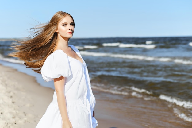 Happy, beautiful woman on the ocean beach standing in a white summer dress. Portrait.