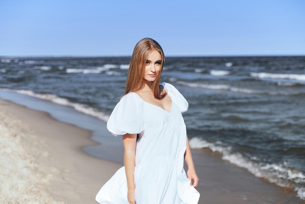 Happy, beautiful woman on the ocean beach standing in a white summer dress. Portrait.