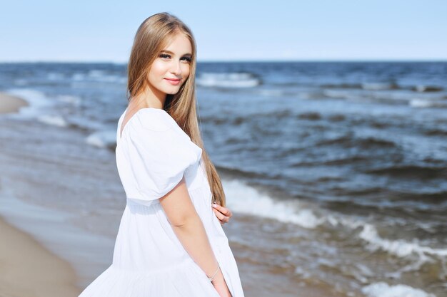 Photo happy, beautiful woman on the ocean beach standing in a white summer dress