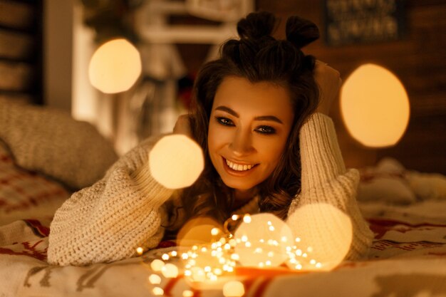 Happy beautiful woman in vintage knitted sweater with festive lights on the bed