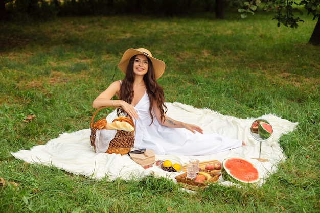 Foto felice bella donna che indossa un cappello a tesa larga e abito bianco in piedi e tenendo il cesto di pane al parco estivo