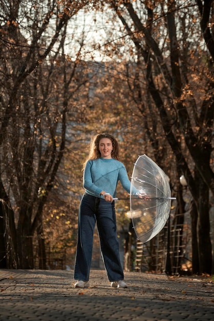 Happy beautiful woman with curly hair dance in alley of autumn park with transparent umbrella Girl walk in park