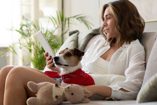 Happy beautiful woman with dog using digital tablet on sofa at home.