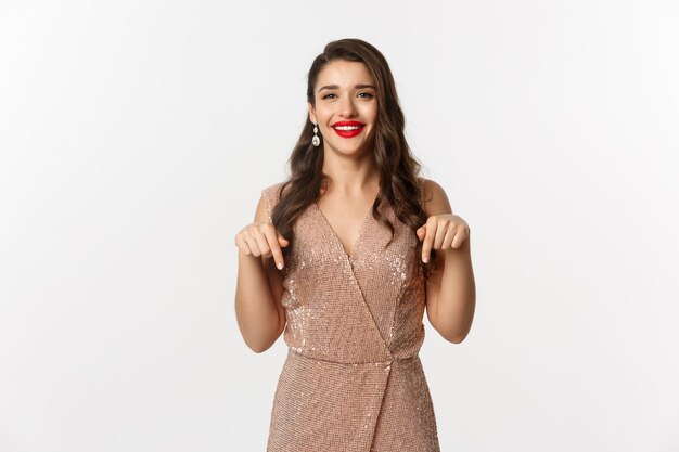 Happy beautiful woman with red lips, smiling while showing promo, wearing elegant dress for christmas party, pointing fingers down, standing over white background