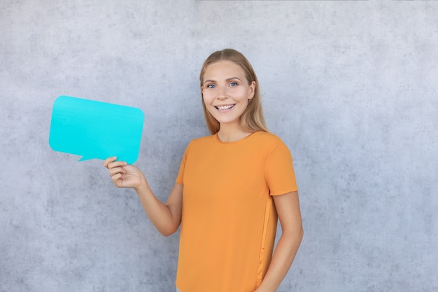 Happy beautiful young blonde woman standing isolated over gray background, holding empty speech bubble.