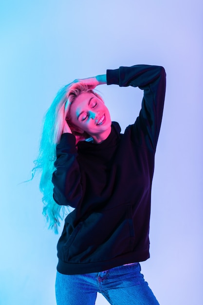Счастливая красивая молодая блондинка с милой улыбкой в модной черной толстовке с синими джинсами на неоновом розовом свете в студии