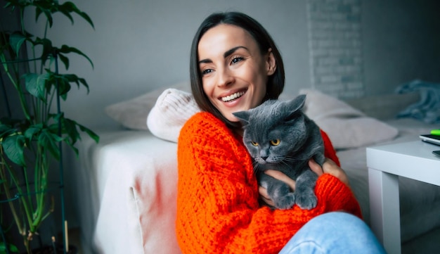 Happy beautiful young brunette woman holding and hugging her cute British shorthair cat on hands and relaxes on the floor at home