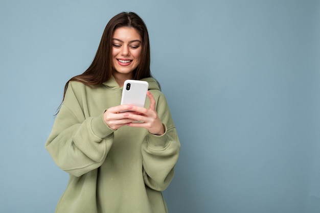 Счастливый красивая молодая брюнетка женщина в стильной зеленой толстовке с капюшоном с помощью мобильного телефона, написание sms, изолированные на фоне, глядя на экран смартфона