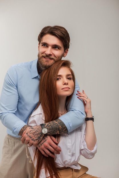Фото Счастливая красивая молодая модная пара красивый хипстер с татуировкой и бородой обнимает красивую рыжую девушку в модной одежде на белом фоне
