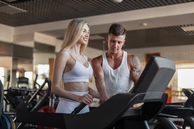 Happy beautiful young fitness girl with a smile in trendy sportswear running on a treadmill in the gym with a fitness instructor guy