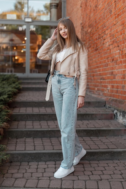 Felice bella ragazza in abiti alla moda con una giacca di pelle, jeans vintage a vita alta e scarpe bianche cammina giù per i gradini della città stile casual urbano femminile e moda