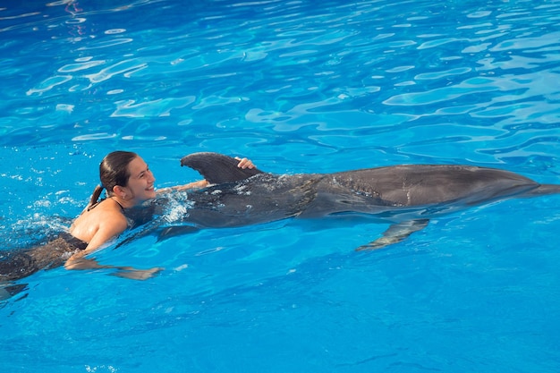 Happy beautiful young girl laughs and swims with dolphins in blue swimming pool on sunny day