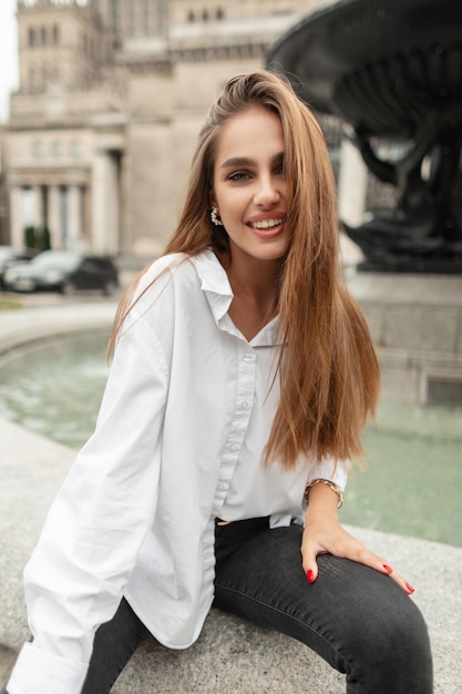 白いシャツと黒いジーンズを着たファッショナブルなカジュアルな服装で笑顔で幸せな美しい若い女の子は、ヨーロッパの町のヴィンテージの噴水の近くに座っています。