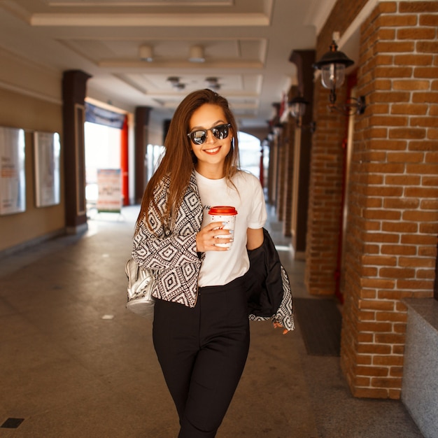 Happy beautiful young model girl with coffee in fashion clothes walking in the city