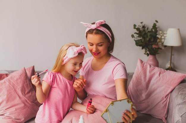 幸せな美しい若い母親と彼女の愛らしい娘の両方がピンクのTシャツとスタイリッシュな柔らかいヘアバンドで日当たりの良いリビングルームの灰色のソファに座って手鏡を見ています