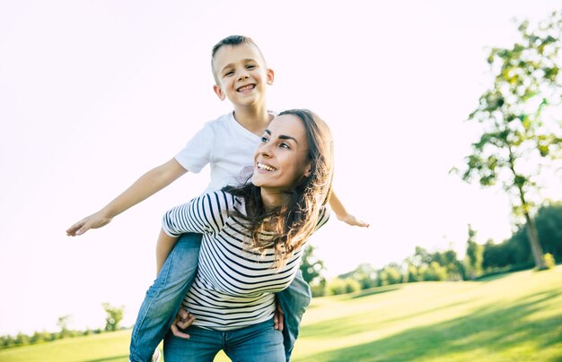 Happy beautiful young mother is playing with her little son while he is riding on her back and shouting for joy