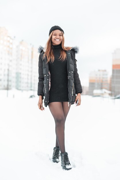 Happy beautiful young sexy african woman in fashionable winter clothes with hat, jacket, sweater, tights and boots walking on the street with snow