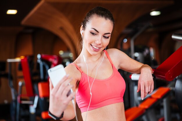 Happy beautiful young sportswoman listening to music and making selfie with smartphone in gym