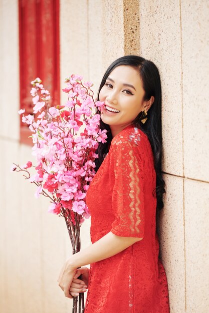 Happy beautiful young Vietnamese woman in red dress standing outdoors with brahches of pink peach blooming branches