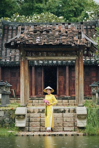 Happy beautiful young Vietnamese woman in yellow ao dai dress and traditional conical hat standing at entrance of wooden temple