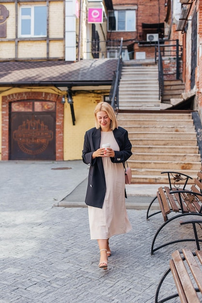 Happy beautiful young woman in dress and jacket walks around the city and drinks coffee