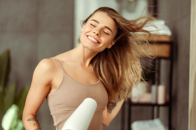 Photo happy beautiful young woman drying her hair with dryer wearing tank top standing in bathroom