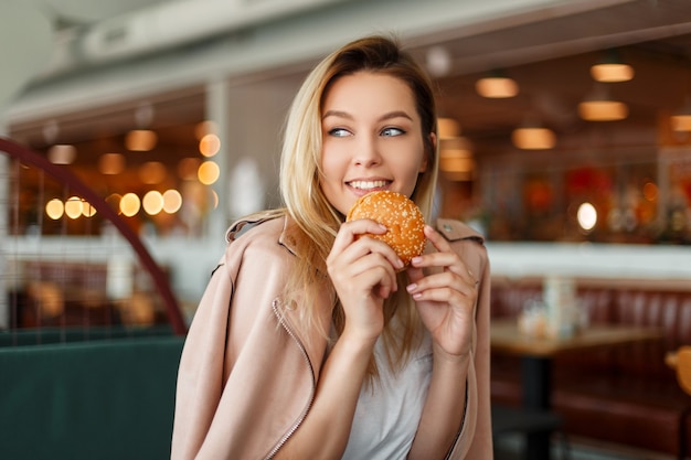 Счастливая красивая молодая женщина ест гамбургер в помещении