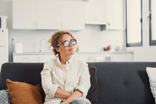 Happy beautiful young woman enjoying leisure at cozy home sitting on new comfortable couch looking away smiling laughing Candid portrait of female model in modern apartmentxA