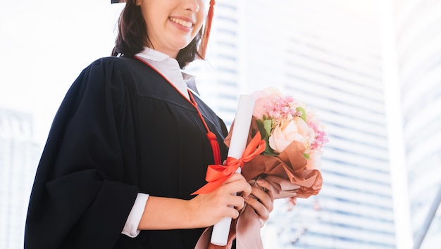 美しい若い女性の卒業生の卒業証書と花の花束を保持している