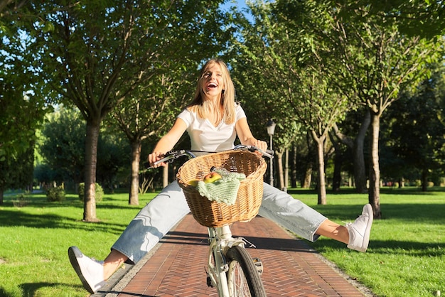 Happy beautiful young woman having fun on retro bike with basket, teenager girl in park raised legs to the sides, sunny summer day in nature