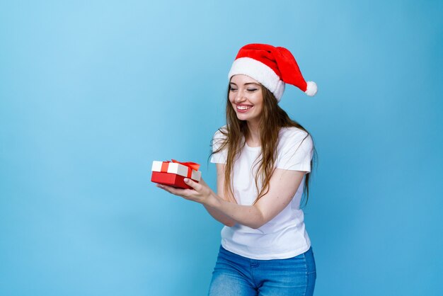 Happy beautiful young woman holding gift box in santa hat and white tshirt on a blue background smil...