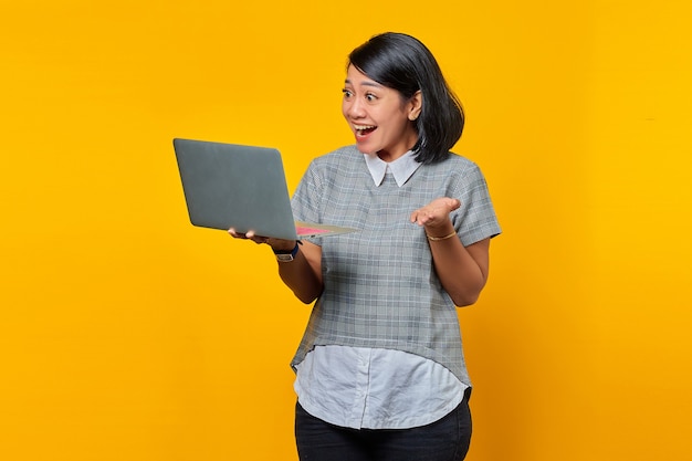 Happy beautiful young woman looking at laptop and raising hand on yellow background