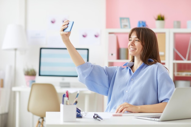 Happy beautiful young woman in oversize blouse sitting at table and taking selfie in office first wo