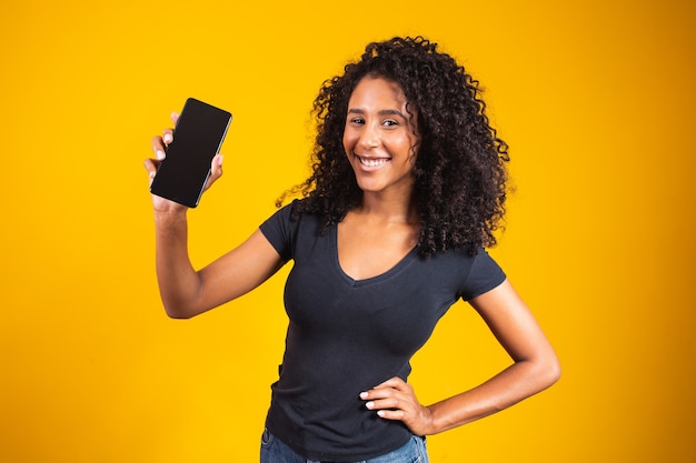 Happy beautiful young woman with afro hair holding blank screen cell phone on yellow background.