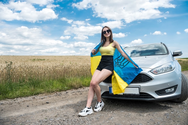 Happy beautiful young woman with flag of ukraine standing near her car at summer road