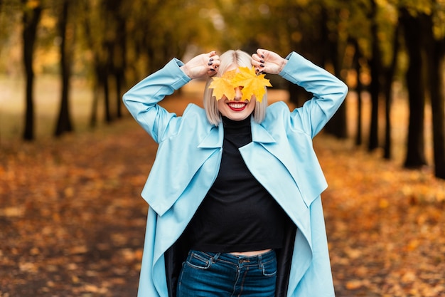 Happy beautiful young woman with a smile in a fashionable blue coat and a black sweater covers her eyes with yellow fall leaves in an autumn park