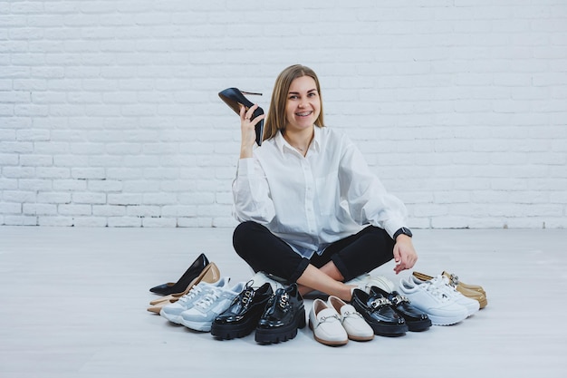 Happy beautiful young woman with a smile in a fashionable outfit with a white shirt and black trousers sits on the floor among the shoes and chooses a new pair of shoes for herself