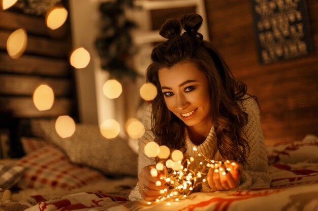 Happy beautiful young woman with a smile in a vintage knitted sweater with holiday lights on the bed