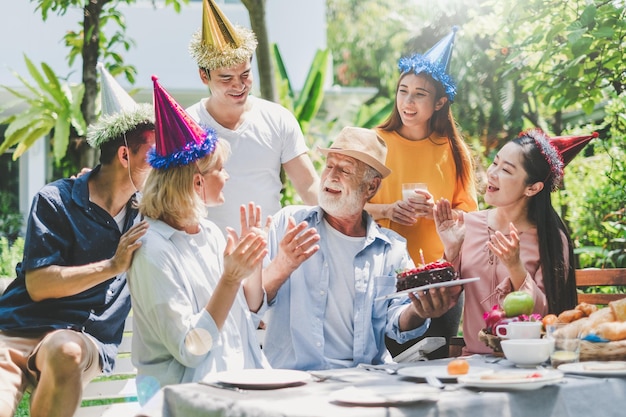 Happy big family have a lunch at outdoor in green garden Grand father's birthday party on picnic table in summer Old man hold the cake Big family outdoor lunch party concept