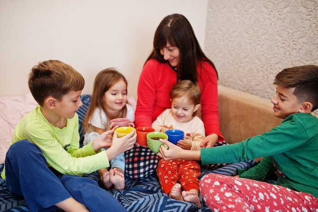 Happy big family is having fun together in bedroom. Large family morning concept. Mother with four kids wear pajamas drink tea in bed at home.