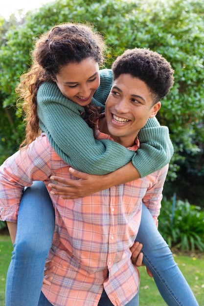 Happy biracial couple having fun piggybacking in garden and smiling. Inclusivity, domestic life, leisure time, romance and togetherness concept.