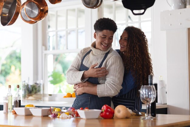 happy biracial couple preparing food, smiling and embracing in kitchen. inclusivity, domestic life, leisure time and togetherness concept.