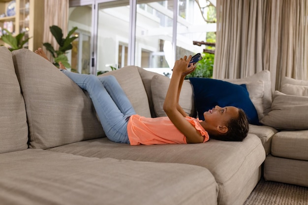 Фото Счастливая девушка двух рас, лежащая на диване и использующая планшет в гостиной. детство, свободное время, технологии, общение, развлечения, образ жизни и домашняя жизнь неизменны.