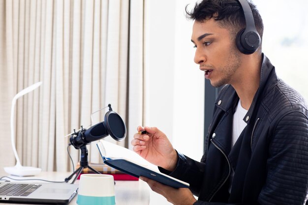 Happy biracial transgender man in headphones, making notes and talking to microphone, podcasting