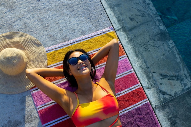 Happy biracial woman lying on towel and sunbathing next to swimming pool in garden. Lifestyle, free time and vacation, summer, sunshine, unaltered.